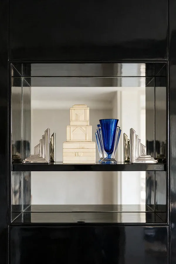 A glossy black built-in display shelf holds a small Art Deco-style vignette with mirrored geometric bookends, a stacked cream decorative sculpture, and a cobalt blue glass vase. The arrangement sits against a light neutral background, creating a stark, elegant contrast.
