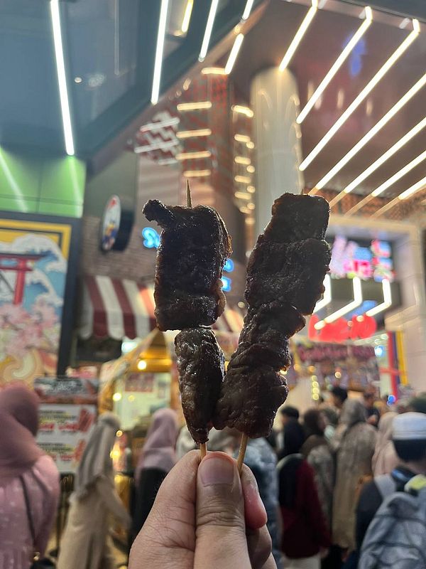 Three skewers of wagyu beef are held up in front of a bustling street scene.