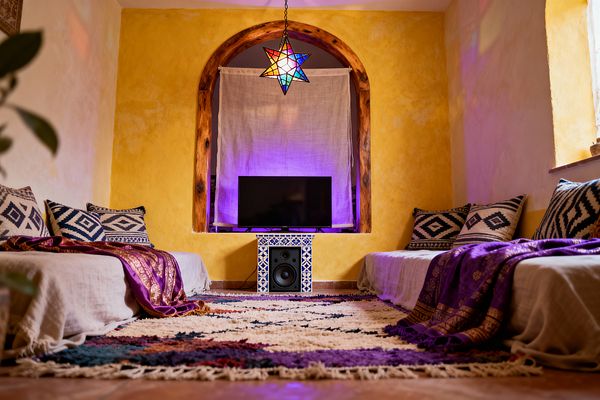 A bohemian media room with two low sofas facing each other, covered in natural linen and layered with indigo-and-ivory patterned cushions and purple, gold-trimmed throws. At the center, an arched niche with rustic wood trim holds a TV on a blue-and-white tiled stand; a linen curtain and violet backlighting sit behind it. A cream, multicolor shag rug spans terracotta floors, while a multicolored stained-glass star pendant casts jewel-toned reflections across sunny yellow plaster walls and a small side window.