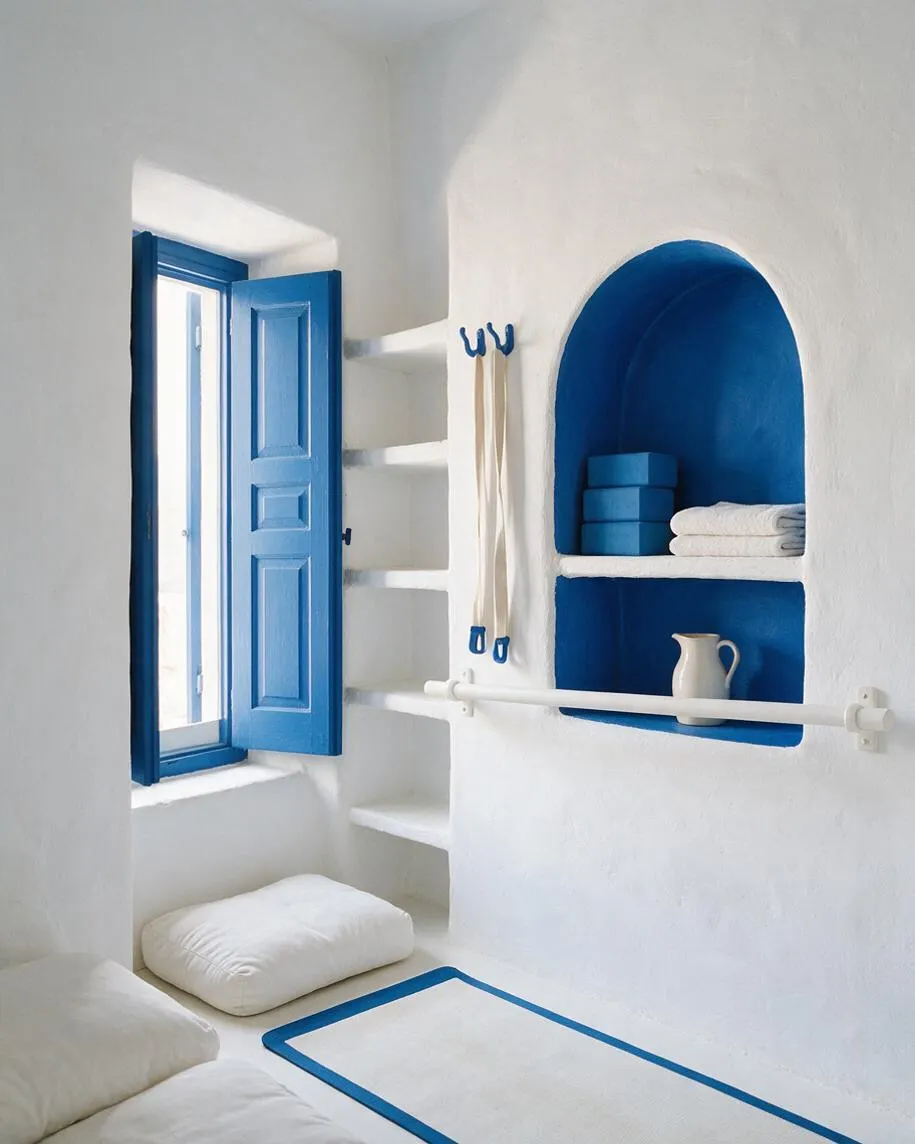A compact home gym with white plaster walls, a bright blue arched niche, and matching blue window shutters. Built-in shelves hold yoga blocks and folded towels, while cushions and a bordered exercise mat sit on the floor in a sunlit, minimalist Mediterranean-inspired space.