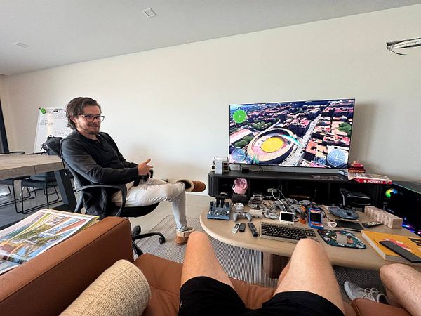 A man sits in a modern workspace with a large TV displaying a bird's-eye view of a stadium, surrounded by various gadgets and a casual atmosphere.