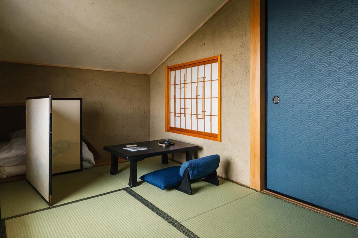 A Japanese Zen guest bedroom with tatami mat flooring, a low black table, and a blue floor cushion. A shoji-style window with wood framing adds warmth, while a folding screen partially separates the sleeping area on the left. A bold blue sliding door with wave pattern brings texture and color to the calm neutral room.