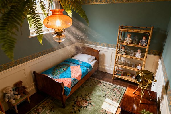 A Victorian-inspired children’s bedroom with a single mahogany sleigh bed covered by a teal and coral quilt featuring a peacock feather motif. Teal walls with a gold floral border sit above white wainscoting, while an amber glass pendant casts sunlit beams near a window and ferns. A bamboo-style shelf displays vintage dolls and glass jars, and a brass magic-lantern projector rests on a polished wood table beside a sage green floral rug and dark hardwood floor.