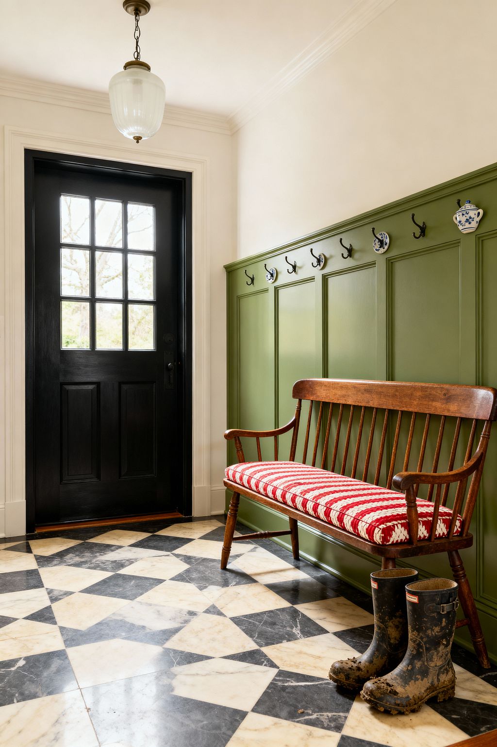 A traditional mudroom features a black, divided-light exterior door, cream walls, and olive green paneled wainscoting fitted with black metal hooks and blue-and-white ceramic pegs. A stained wood spindle-back bench with a red-and-white crocheted cushion lines the wall above a black-and-cream harlequin marble floor, while a milk-glass pendant hangs overhead and a pair of muddy rubber boots rest near the bench, signaling the space’s durable, welcoming purpose.