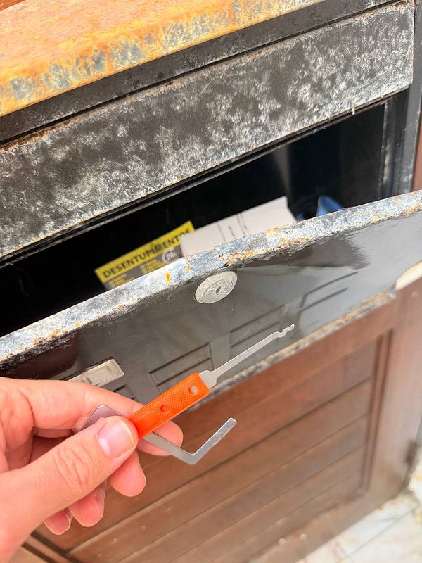 An open rusty metal mailbox with a hand holding an orange lock pick tool near the lock.