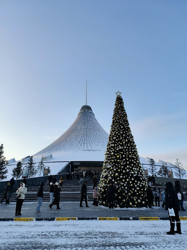 A winter scene at Khan Shatyr Mall featuring a large Christmas tree and visitors.