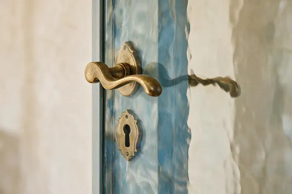 A close-up detail of a French Country style door showing a curved antique brass handle and matching keyhole escutcheon mounted on textured, frosted glass. The softly blurred glass and warm metal hardware create a refined, old-world feel against a pale neutral background.