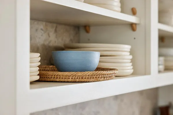 A close-up of open white kitchen shelving styled with stacks of cream-colored plates, a pale blue bowl, and a woven rattan tray. The arrangement sits against a soft neutral backsplash, creating a light and relaxed coastal look.