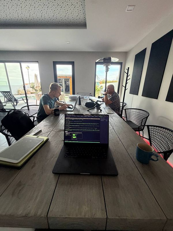 Two men working at a long wooden table with laptops in a modern coworking space with outdoor views.
