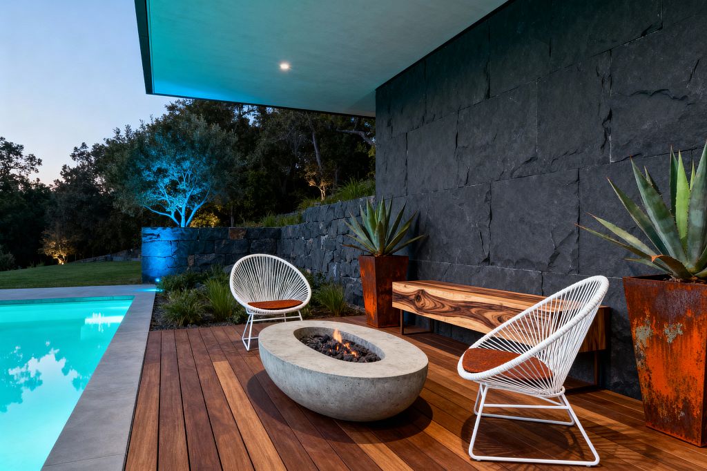 Outdoor patio by a pool featuring an ipe wood deck and a charcoal slate wall. At the center sits a low oval cast-concrete fire pit, flanked by two white Acapulco-style chairs with rust-colored cushions and a floating live-edge acacia bench. Two tall corten steel planters hold spiky agave. A flat roof soffit with a recessed light extends overhead, while blue and warm uplights illuminate trees beyond the stone retaining wall.