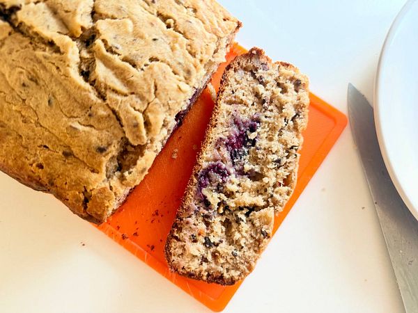 A freshly baked banana cake loaf with visible blueberries and cacao nibs, sliced on an orange cutting board.