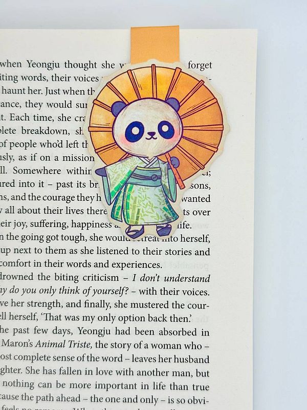 A colorful panda bookmark is placed on a page of a book, featuring a character in traditional attire.
