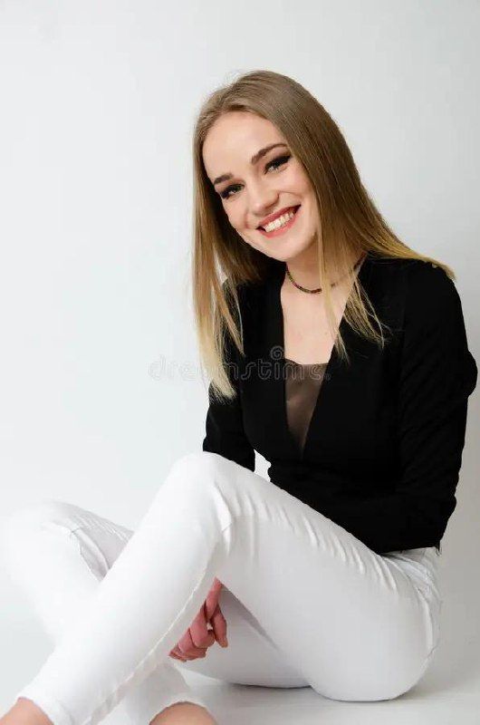 A young woman with straight blonde hair sits smiling against a plain white background, wearing a black top and white pants.