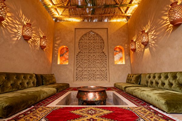 A cozy Moroccan media room with low olive green velvet bench seating lining two walls around a sunken center that holds a hammered copper bowl table. Sand-colored tadelakt walls feature a carved pointed-arch geometric screen and two arched niches backlit with veined onyx, while terracotta perforated sconces throw patterned shadows and a reed-thatched ceiling glows warmly above layered red kilim rugs.