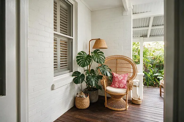A tropical front porch with white painted brick walls, a dark wood floor, and a woven rattan peacock chair accented by a pink floral cushion. A tall potted monstera, woven basket, and woven floor lamp create a relaxed, layered feel beside shuttered windows and an open view to lush greenery outside.