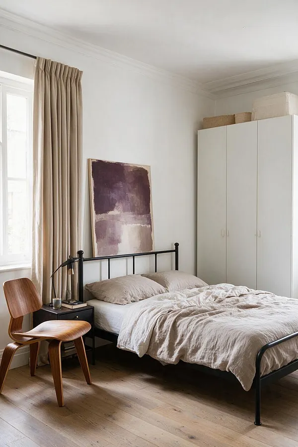 A modern master bedroom with a black metal bed frame, beige bedding, and a simple abstract painting leaning against the wall. Soft beige curtains, a wood chair, and a white wardrobe create a calm, minimalist look with natural light and warm wood floors.