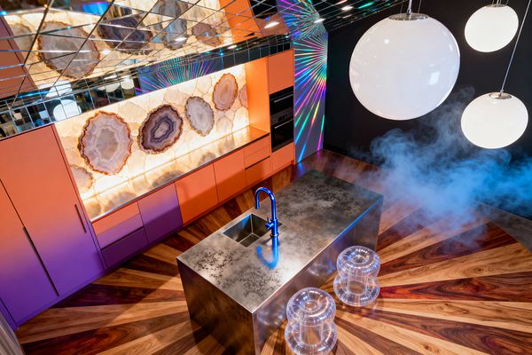 A contemporary kitchen with orange-to-purple gradient cabinets, a backlit agate stone backsplash, and a patinated stainless-steel island with a blue faucet. A mirrored tile ceiling, opal globe pendants, clear acrylic stools, and a radial walnut sunburst floor amplify rainbow light projections on a black wall, creating a surreal, high-gloss atmosphere with built-in black appliances.