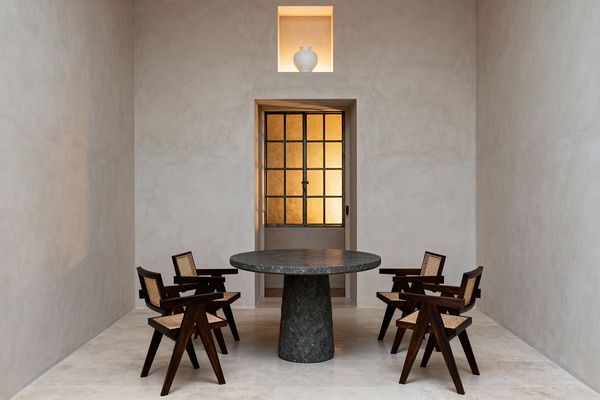 A minimalist dining room with a round charcoal stone pedestal table surrounded by four dark wood armchairs with woven cane seats and backs. The space features warm white plaster walls and pale limestone flooring, a steel-framed door with amber textured glass glowing from behind, and a small illuminated wall niche above the doorway displaying a white ceramic vase, all arranged symmetrically in a narrow, high-ceilinged room with calm, even lighting.