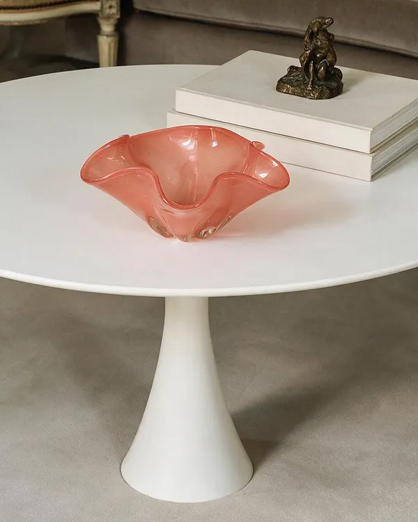 A round white pedestal coffee table holds a coral-pink handblown glass bowl and a stack of white books topped with a small bronze sculpture. The setting feels refined and eclectic, with soft neutral carpeting and muted furniture in the background.