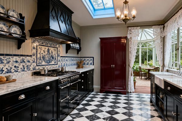 A Victorian-style galley kitchen with glossy black cabinets and chrome hardware features white marble countertops and a black-and-white checkerboard marble floor. The backsplash is blue-and-white Delft tile with a central panel beneath a carved black hood above a large range. A brass chandelier and a ceiling skylight illuminate the room. On the right, an apron-front sink faces tall windows dressed in lace curtains that open to a glass conservatory. A deep burgundy freestanding pantry stands against a beadboard wall.