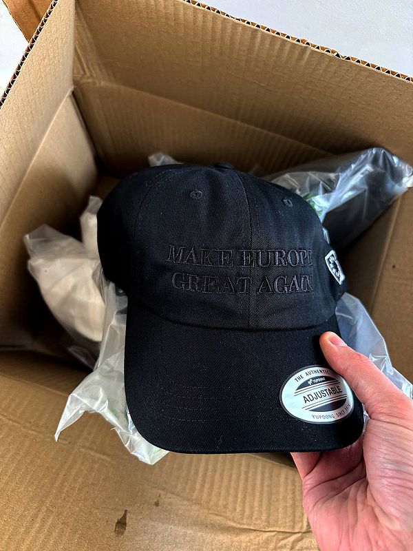 A black cap with embroidered text is being held above a cardboard box containing merchandise.