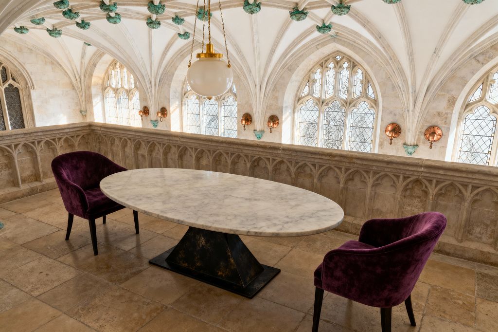 A compact dining vignette set in a Gothic stone gallery with ribbed vaults and pointed arched, leaded-glass windows. An oval white marble table on a blackened pyramidal base is paired with two deep purple velvet armchairs; a brass-chained globe pendant and round copper wall sconces warm the honey-beige limestone floors, carved tracery balustrade, and verdigris ceiling bosses.