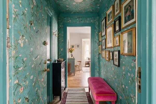 A narrow entryway with turquoise floral wallpaper covering the walls and ceiling, framed artwork arranged salon-style on the right wall, and brass door hardware on the left. A bright pink velvet bench with gold trim sits along the hallway beside layered runners, leading to a light-filled room beyond.