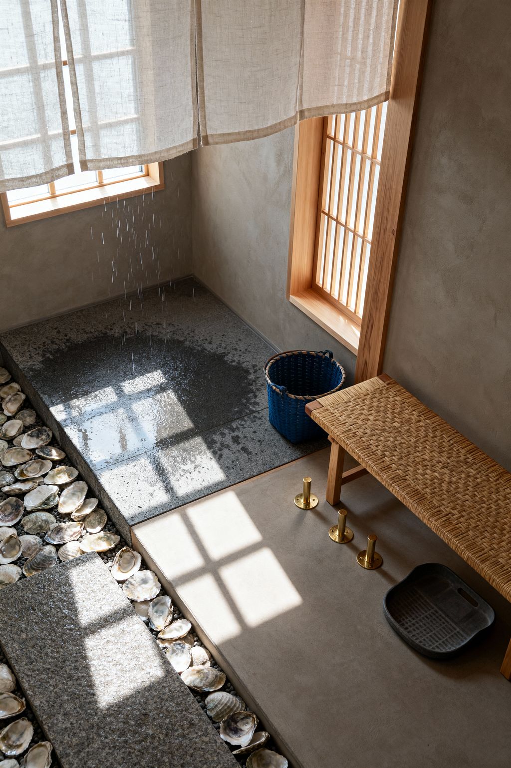 A serene Japanese-style mudroom features a raised plaster platform with a woven rattan bench, three floor-mounted brass pegs, and a dark rubber boot tray. Beside it, an indigo basket sits near a lower granite slab where water drips to rinse footwear, bordered by a channel filled with oyster shells and a textured stone tread. Light wood windows with slatted screens and a linen curtain filter soft natural light across the warm, minimal space.
