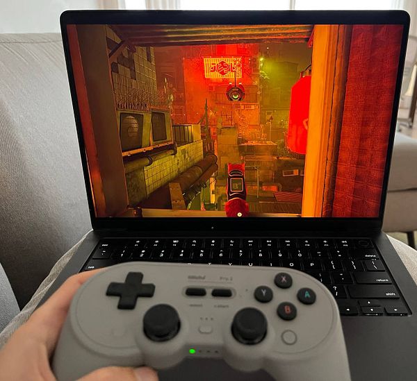 A gaming setup featuring a MacBook Air with an 8BitDo Pro 2 controller connected, a cup of coffee, and Stray game artwork on the screen.