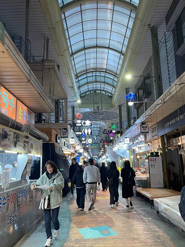 A bustling market scene with people walking through a covered walkway lined with food stalls.