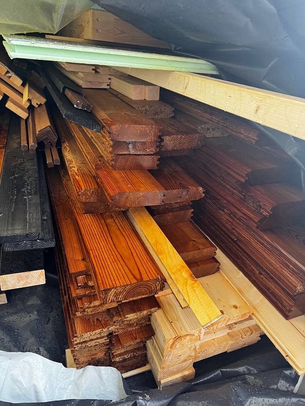 A stack of various wooden planks is seen covered with a tarp in a garden setting.