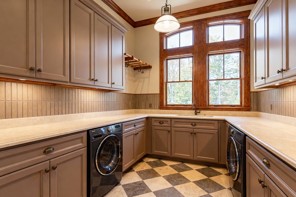 A transitional laundry room with U-shaped countertops and taupe Shaker cabinets, lit by under-cabinet lighting and a central dome pendant. Beige stone-look counters and a vertical rectangular tile backsplash wrap the room; two graphite front-loading machines sit beneath the worktops. A small undermount sink is centered under large arched windows framed in warm stained wood. The floor shows a charcoal-and-cream checkerboard pattern, creating a classic yet streamlined workspace.