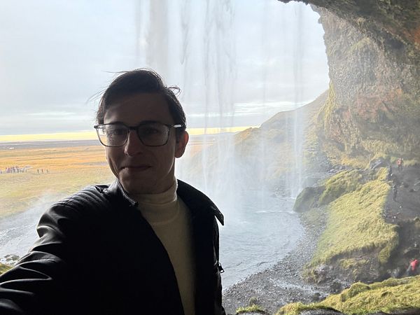 A person stands in front of a waterfall, capturing a selfie with a scenic backdrop.