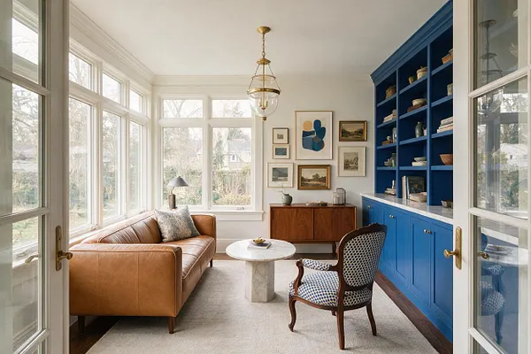 A light-filled sunroom with floor-to-ceiling windows, French doors, and a camel leather sofa beside a small marble coffee table. Bright cobalt blue built-in shelves and cabinets line one wall, balanced by framed artwork, a patterned accent chair, and warm wood furniture.