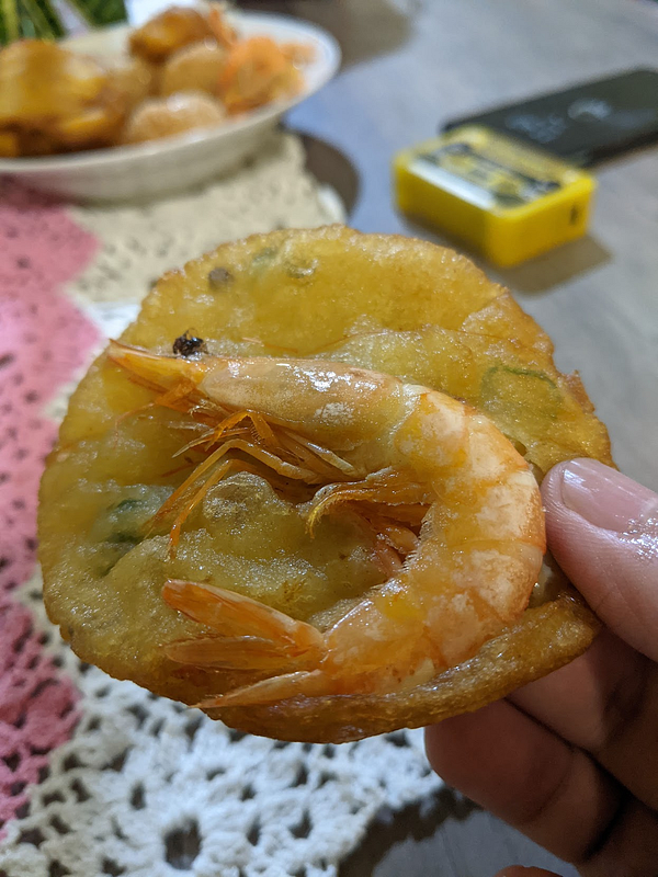 A hand holds a shrimp fritter, showcasing its crispy texture and a shrimp on top.