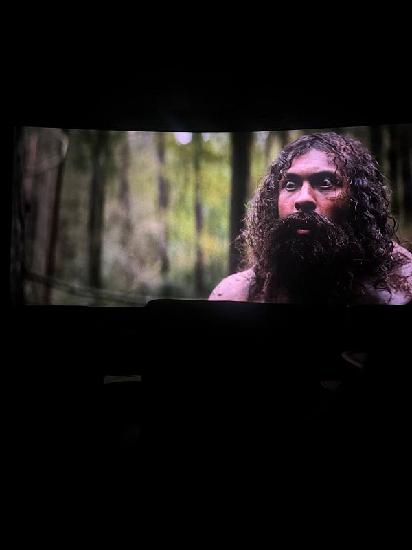 Portrait photo of a young woman with long blonde hair smiling, leaning on a wooden surface against an ivy-covered brick wall.