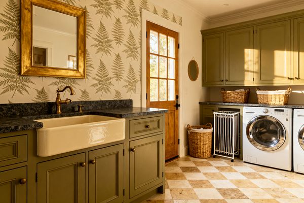 A traditional laundry room features olive green inset cabinets with bronze knobs, a cream apron-front farmhouse sink, and dark stone countertops. Fern-patterned wallpaper, a gilded rectangular mirror, and a natural wood glass-panel door add warmth, while a beige-and-cream checkerboard stone floor, front-loading washer and dryer with chrome trim, wicker baskets, and a white rolling laundry sorter complete the organized, classic space.