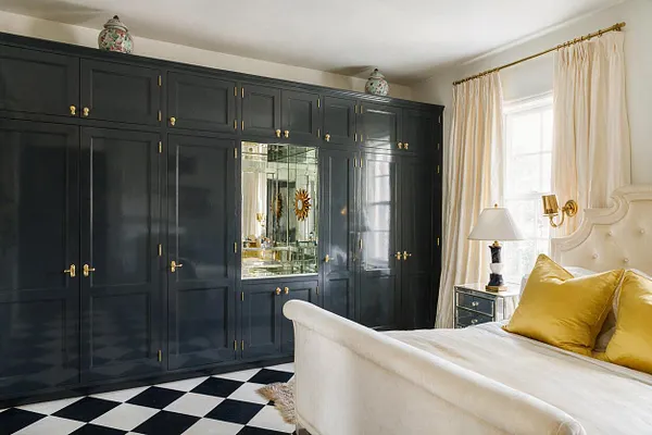 A glamorous master bedroom features a wall of glossy charcoal built-in wardrobes with brass hardware and a mirrored center cabinet. A cream upholstered bed with gold pillows sits beside a black nightstand, while sheer ivory curtains soften the bright window. The black-and-white checkered floor adds a dramatic Hollywood Regency feel.
