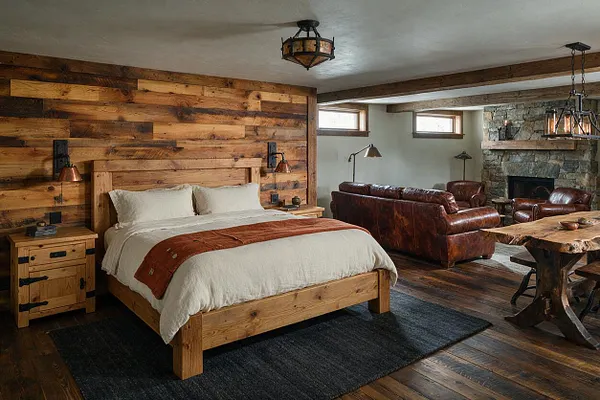 A rustic basement combines a cozy bedroom and lounge area. The room features a wood plank accent wall behind a wooden bed, leather seating around a stone fireplace, exposed beams, and a live-edge wood table under warm lantern-style lighting.