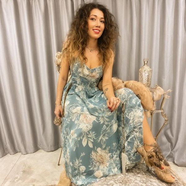 A woman with curly hair is seated in a floral dress against a backdrop of gray curtains.