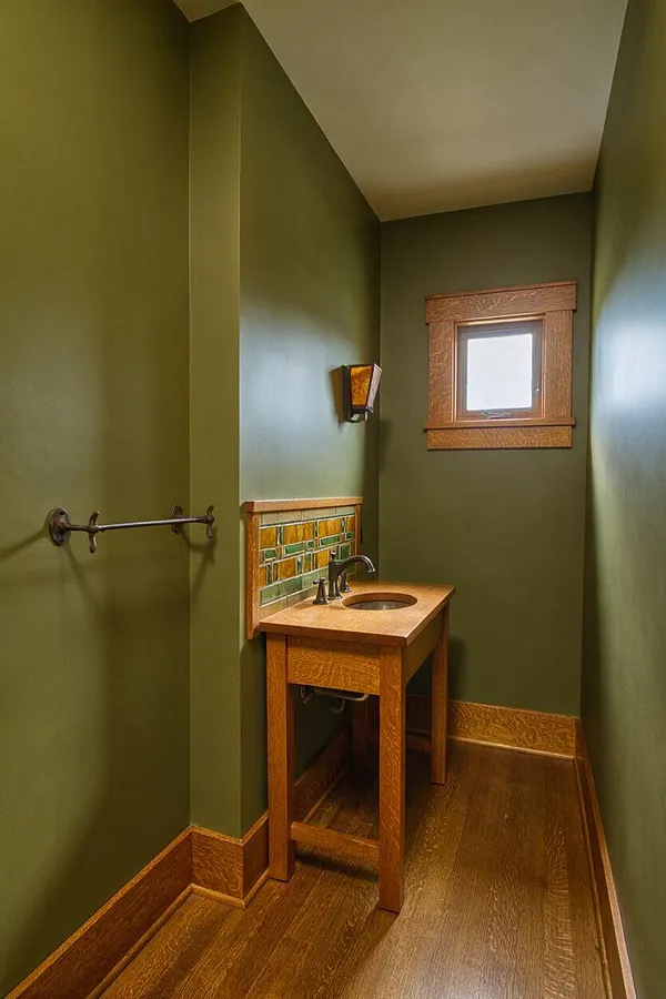 A narrow Craftsman powder room with olive green walls, warm wood trim, and a small wooden vanity with a round sink and dark faucet. A stained-glass-style wall sconce, a small window with a wood frame, and a tiled backsplash add character to the compact space.