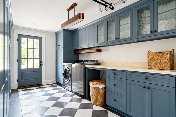 Transitional laundry room featuring dusty blue Shaker cabinets with textured-glass uppers, matte black hardware, and a long light beige stone countertop; a stainless washer and dryer sit beneath a walnut floating shelf. The room is grounded by a diagonal charcoal-and-white checkerboard tile floor and brightened by a multi-lite blue exterior door, white walls, and a slim bronze linear pendant overhead.