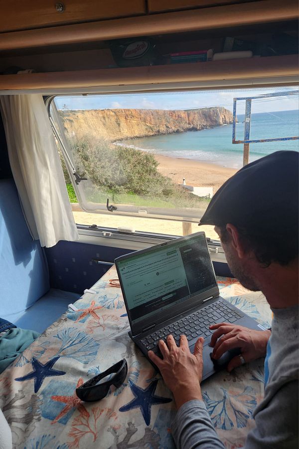 A person is working on a laptop inside a camper van with a scenic beach view.