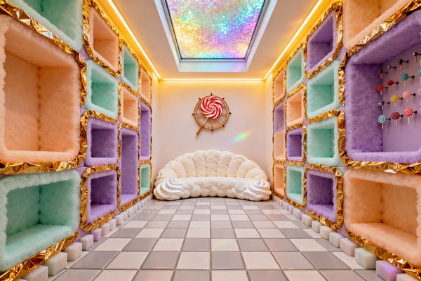A narrow mudroom features floor-to-ceiling built-in cubbies on both sides, each lined in fluffy pastel fabric—mint, peach, and lavender—with metallic gold foil edges. At the end, a scalloped white sofa shaped like whipped cream sits under a round lollipop wall accent, while a glossy gray-and-white tiled floor and warm cove lighting reflect color from a rainbow-iridescent skylight overhead, creating a candy-themed, playful atmosphere.