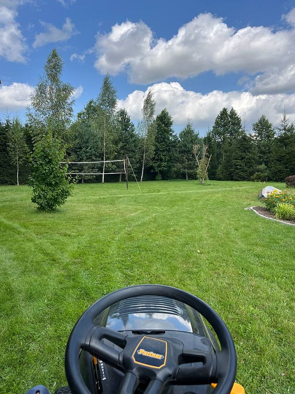 A view from a riding lawn mower in a lush green yard under a partly cloudy sky.