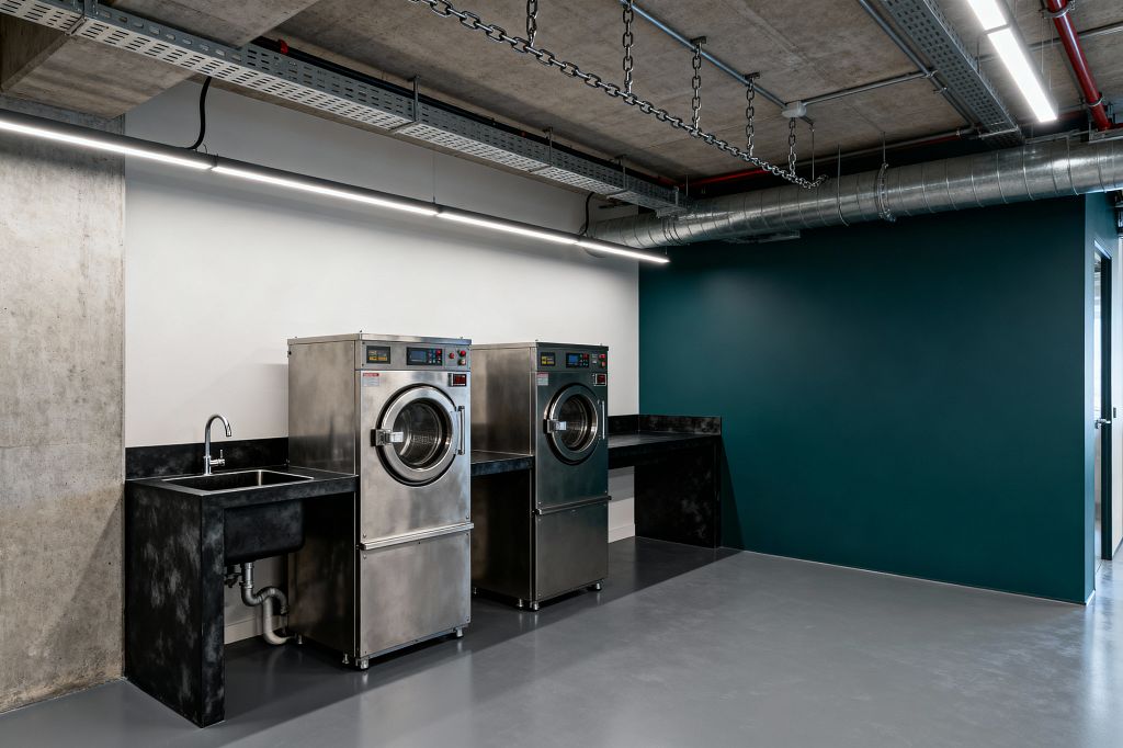 A minimalist industrial laundry room features two stainless-steel front-loading commercial washers between a deep utility sink and blackened steel counters. Surfaces include a seamless gray epoxy floor, a white wall behind the machines and a deep teal accent wall, while overhead an exposed concrete ceiling carries linear LED lights, metal cable trays, chains, red pipework, and a galvanized duct; visible under-sink plumbing and a low black backsplash underscore the utilitarian design.