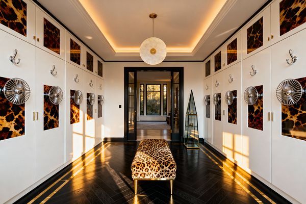 A symmetrical mudroom with tall white cabinets on both sides, detailed with glossy tortoiseshell panels, polished chrome hooks, and circular chrome medallions. The floor is ebonized wood in a herringbone pattern with a brass inlay border. A leopard-print bench with brass legs sits at center. Overhead, an alabaster disc pendant and concealed cove lighting glow. Black-framed glass doors and a triangular glass vitrine complete the space.