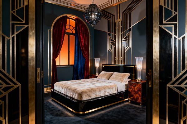 An Art Deco bedroom with deep teal walls overlaid with gold geometric paneling; a black lacquer platform bed with cream faux-fur bedding; burl wood nightstands with tall glass lamps; arched window framed by red and cobalt velvet drapes; a smoked glass pendant hangs above; black and gold double doors and dark carpet; warm under-bed lighting and amber window glow create a dramatic mood.