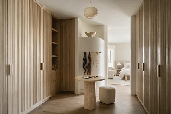 A modern closet hallway with floor-to-ceiling fluted wood cabinetry on both sides, slim vertical brass-toned pulls, and a soft neutral palette. A small stone table and textured round stool sit in the center beneath a simple pendant light, with a bedroom visible at the far end.