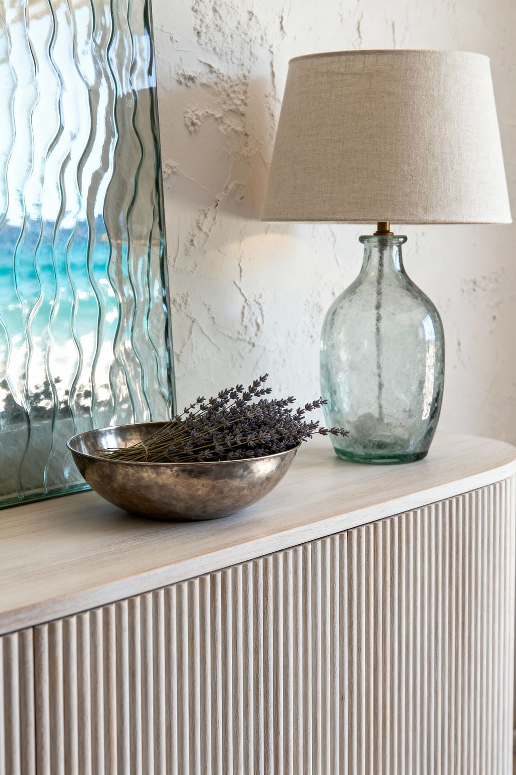 A close-up of a coastal living room vignette shows a whitewashed, fluted-wood console topped with a green-tinted glass bottle lamp with a beige linen shade, a hammered brass bowl holding dried lavender, and a tall rippled glass panel reflecting aqua tones. A sun-washed, textured plaster wall sits behind, with soft natural light enhancing the breezy, beach-inspired mood.